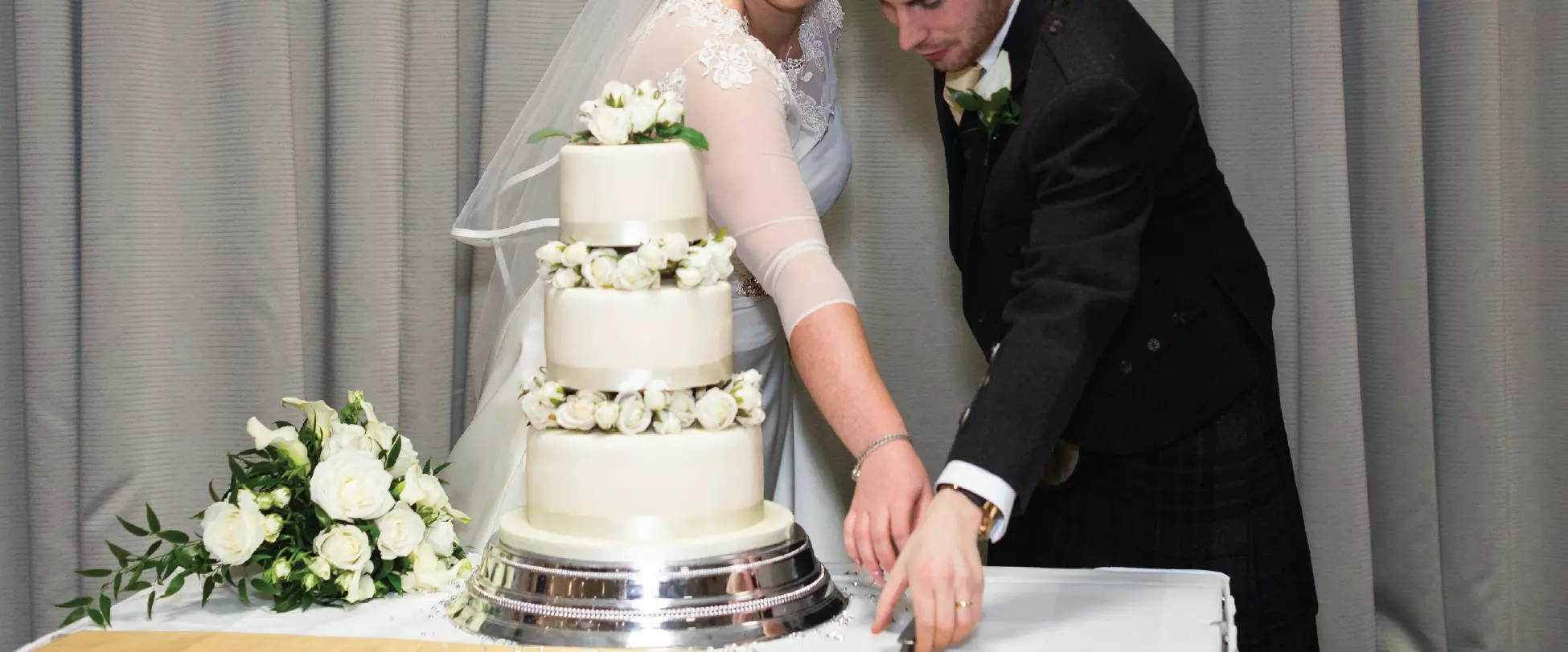 A bride and groom stood behind a table with a 3 tier, white wedding cake on it. There's a bouquet of white flowers to the left hand side of the table. The bride and groom are reaching for a knife to cut the cake..