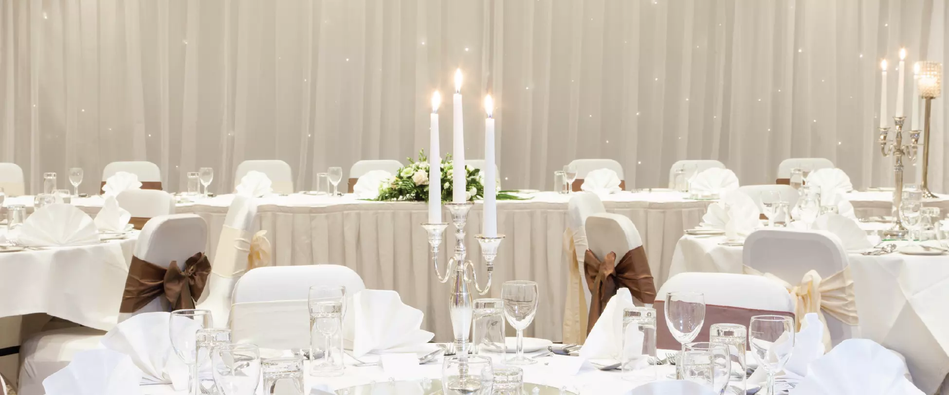 A room with circular tables set up for a wedding breakfast. There's a long table at the back for the bride and groom. The tables are set beautifully with white linens, bows, flowers and large silver candlesticks..